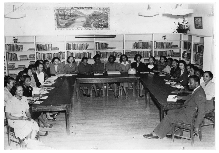 1950~ Blackshear School Faculty Black and white photo of teachers sitting at tables in a U-shaped formation in 1950s period dress. The room has a wood floor and the walls are lined with small bookshelves.